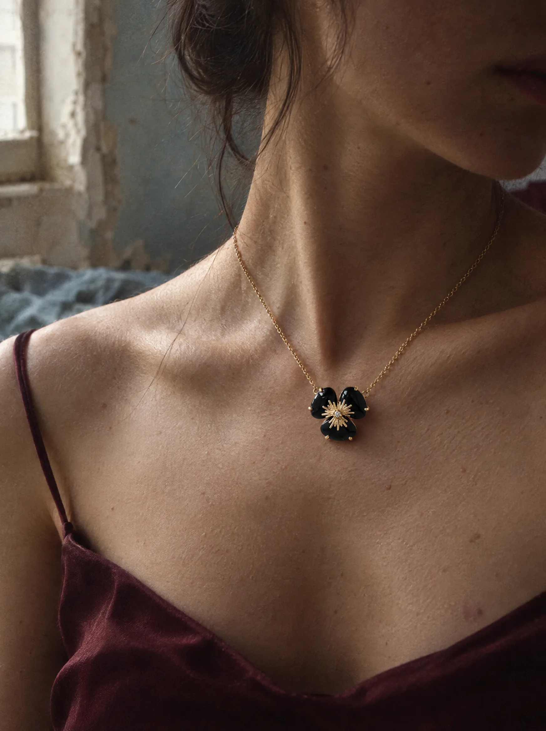 Close crop of a woman's neck and collarbone in a deep wine silk slip, face turned away into shadow, wearing a pendant of three black quartz petals around a gold starburst on a fine gold chain.